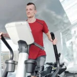 Mies ajaa crosstrainer-kuntopyörällä kuntosalilla keskittyneenä treeniinsä erilaisten kuntosalilaitteiden keskellä.