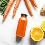 Porkkanoita, inkivääriä ja appelsiinimehua aseteltuna valkoiselle pinnalle esittelemässä inkiväärishotti -valmistetta.